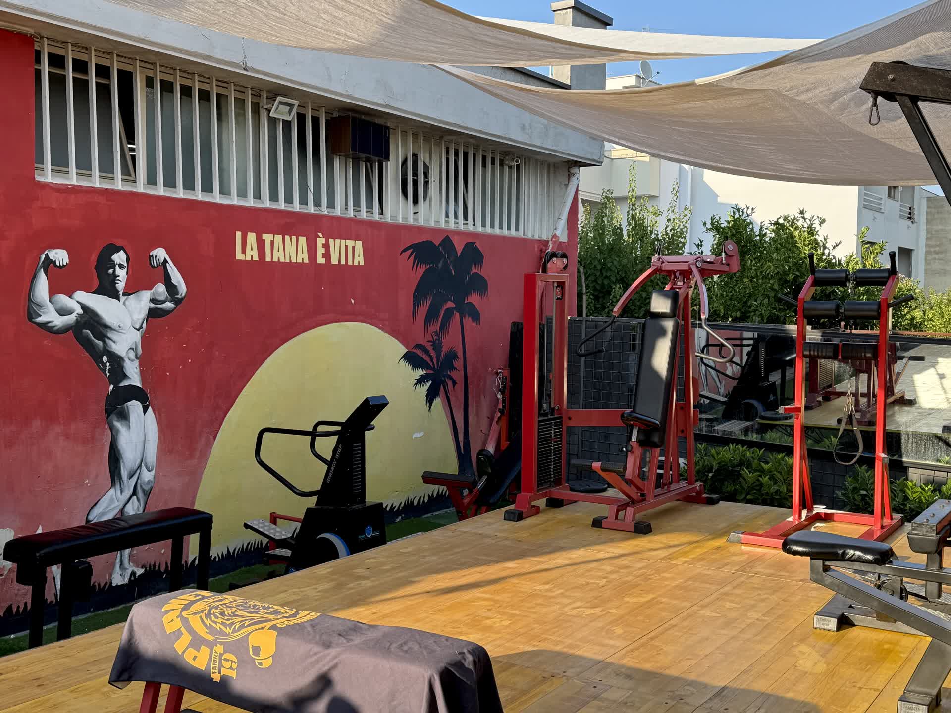 Planet Gym — macchine outdoor davanti al murale La Tana è Vita